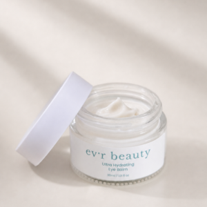Open clear glass jar of ev'r beauty Ultra Hydrating Eye Balm (30ml / 1.01 fl oz) filled with rich white cream, lid leaning against it, labeled in teal script on a white band, set against a soft beige background with gentle natural light and shadow.